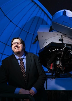 "Peter Plavchan in a suit, seated next to a telescope, with an observatory slit in the background"