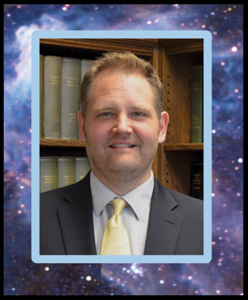 A smiling clean-shaven man in a suit and tie in front of full bookcases