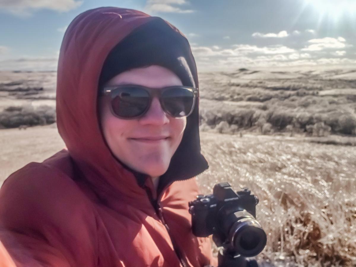 Young man smiling, wearing sunglasses, and holding a camera in a field