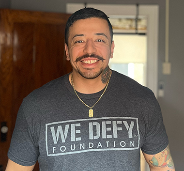A smiling man with short-cropped hair and mustache wearing a shirt that says, WE DEFY foundation. 